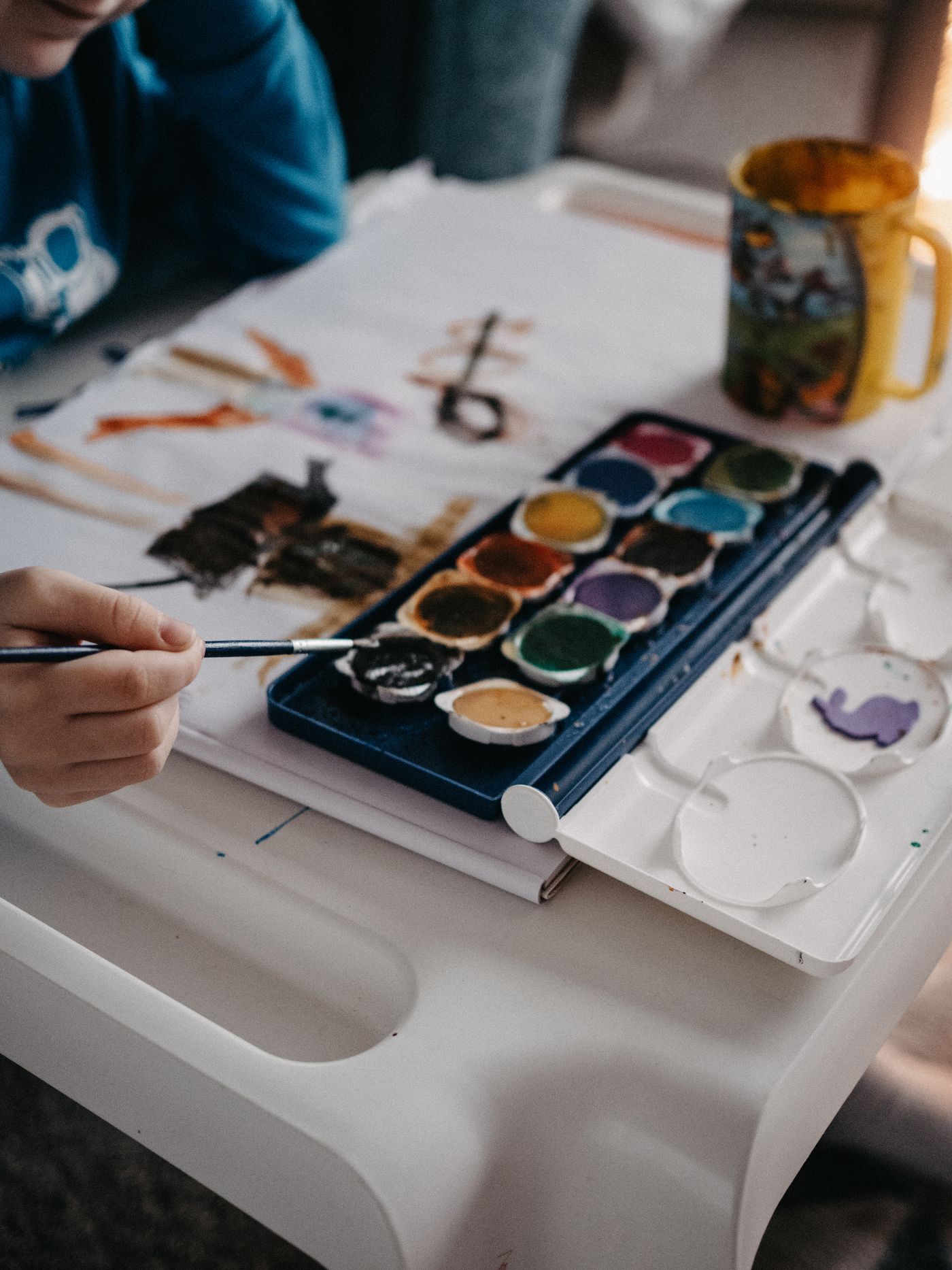 A hand holding a paintbrush beside a watercolour palette and painted paper
