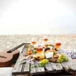 Picnic set up on an old wooden structure on a beach with wine and food. A guitar is off to the side.