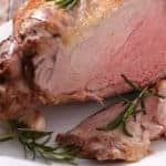 Photo of medium rare leg of lamb cooked perfectly on a white platter with rosemary.