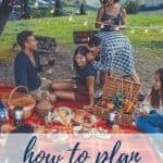 A group of friends sitting on a picnic blanket with food all around. It's dusk and there are white string lights. A man whispers into a woman's ear and she is smiling. A picnic basket with goodies is on the blanket as well.