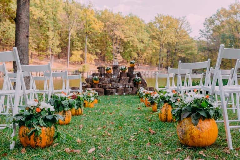 A fall wedding with white folding chairs set up outside with pumpkins at the end of every aisle.