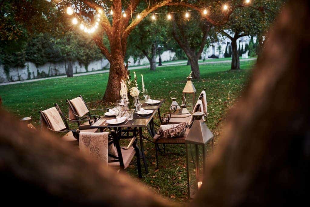 A table set up outside for dining with string lights above, lanterns, simple linens, candlelight, and blankets to keep cozy with dining Al Fresco