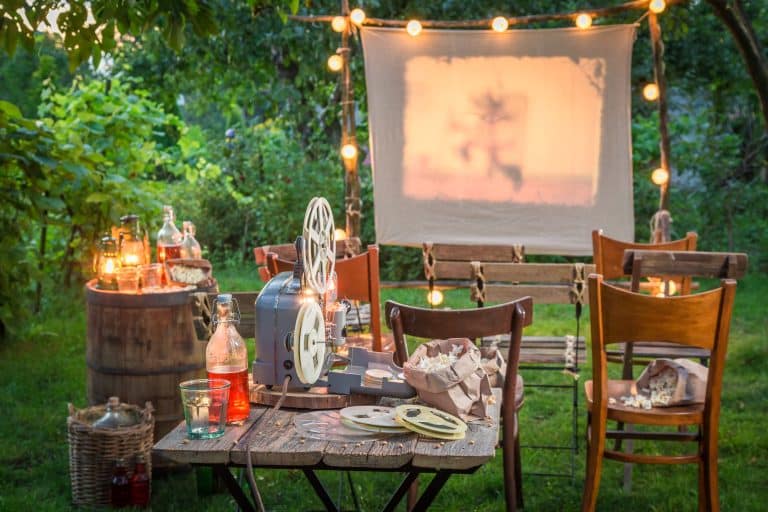 Backyard set up for a movie viewing with mis-matched wooden chairs, a wooden table with old time movie reels, and popcorn. It's evening and there are soft backyard lights.