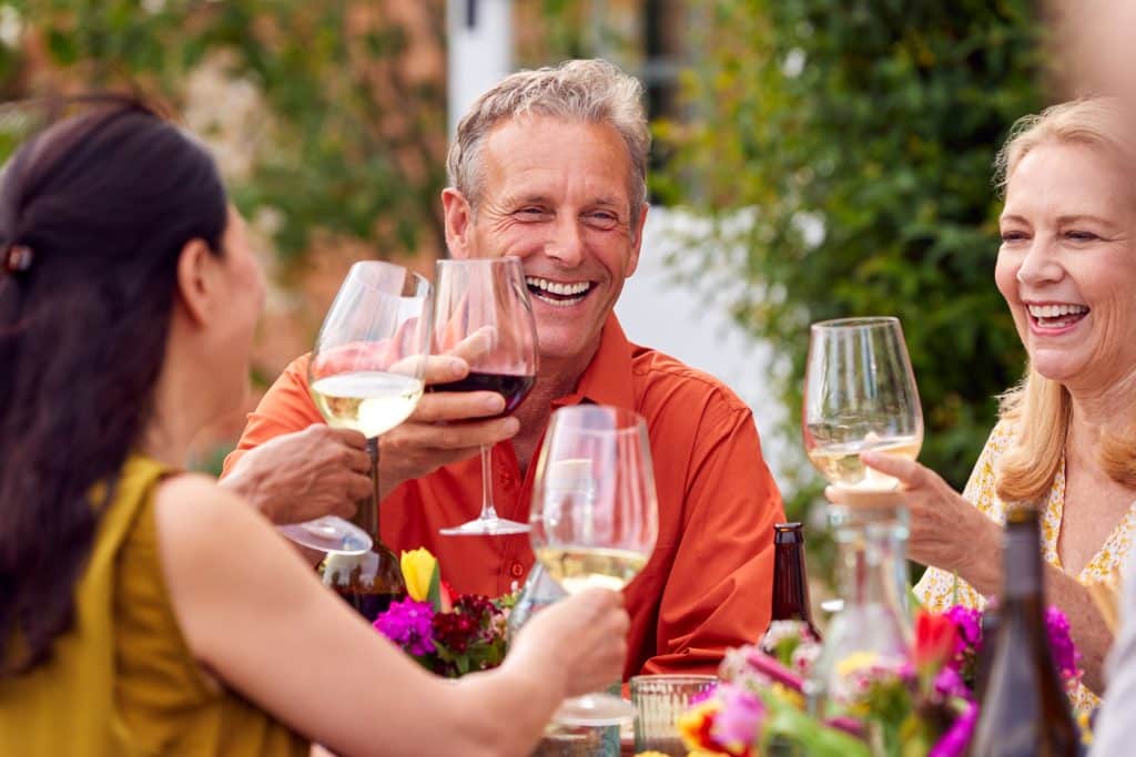 Photo of four people enjoying a glass of wine together, smiling. Three glasses of white and one of red. There are pink and yellow flowers on the table and they are outside in the summer.