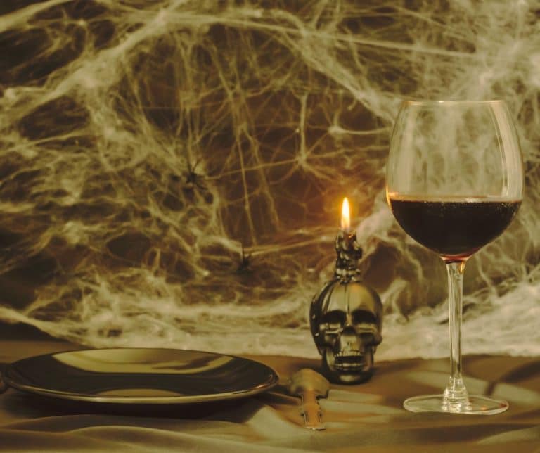 A creepy table setting with cobwebs in the background, dimly lit, dark plate, a silver skull, and a glass of red wine.