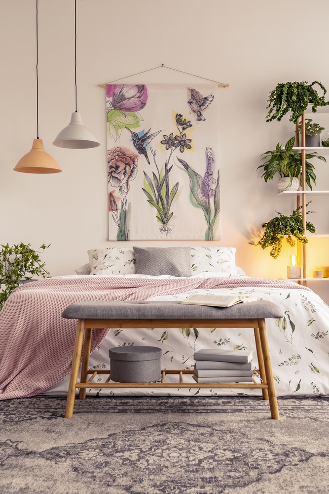 Bench with grey box and books standing by the bed with floral bedclothes and pastel pink blanket in the real photo of bright bedroom interior with fresh plants, lamps and fabric with painted flowers
