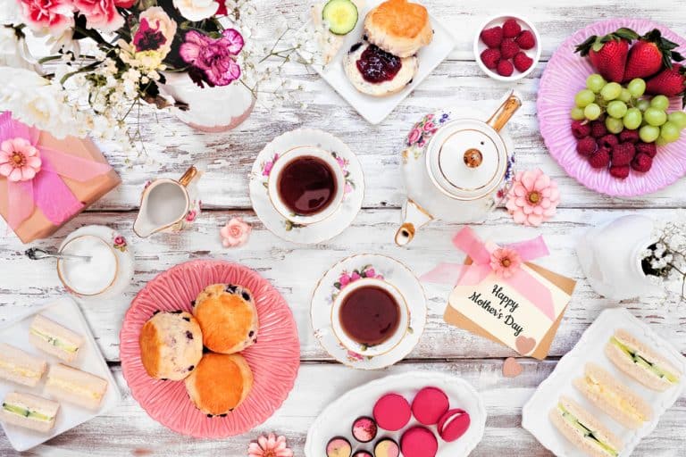 Mother's Day High Tea table scene over a white wood background. Greeting card, gift, flowers, vintage tea set, finger sandwiches, chocolates, desserts and fruit. Sweet Humble Home