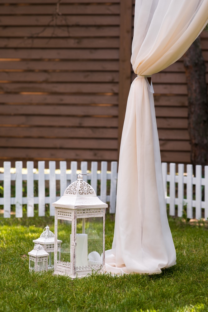 Beautiful cocktail wedding reception outdoors. A white picket fence surrounds a yard of plush, green grass. Old fashioned white hurricane lanterns sit on the ground next to the side of a white tent.