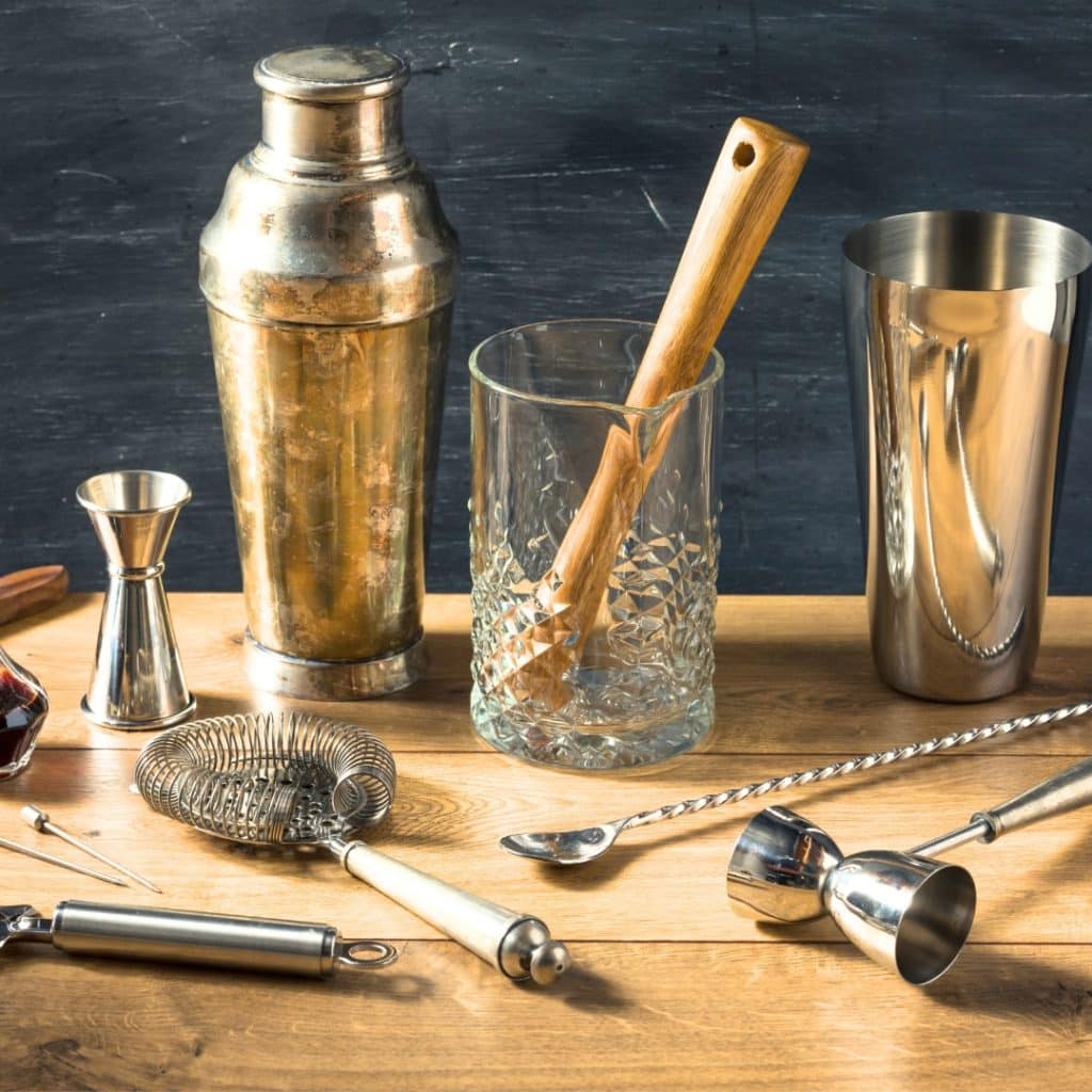 Home Mocktail Bar tools set on a wooden table. Tools include shakers, strainers, jiggers, spoons, a small glass pitcher, and a mixing cup.